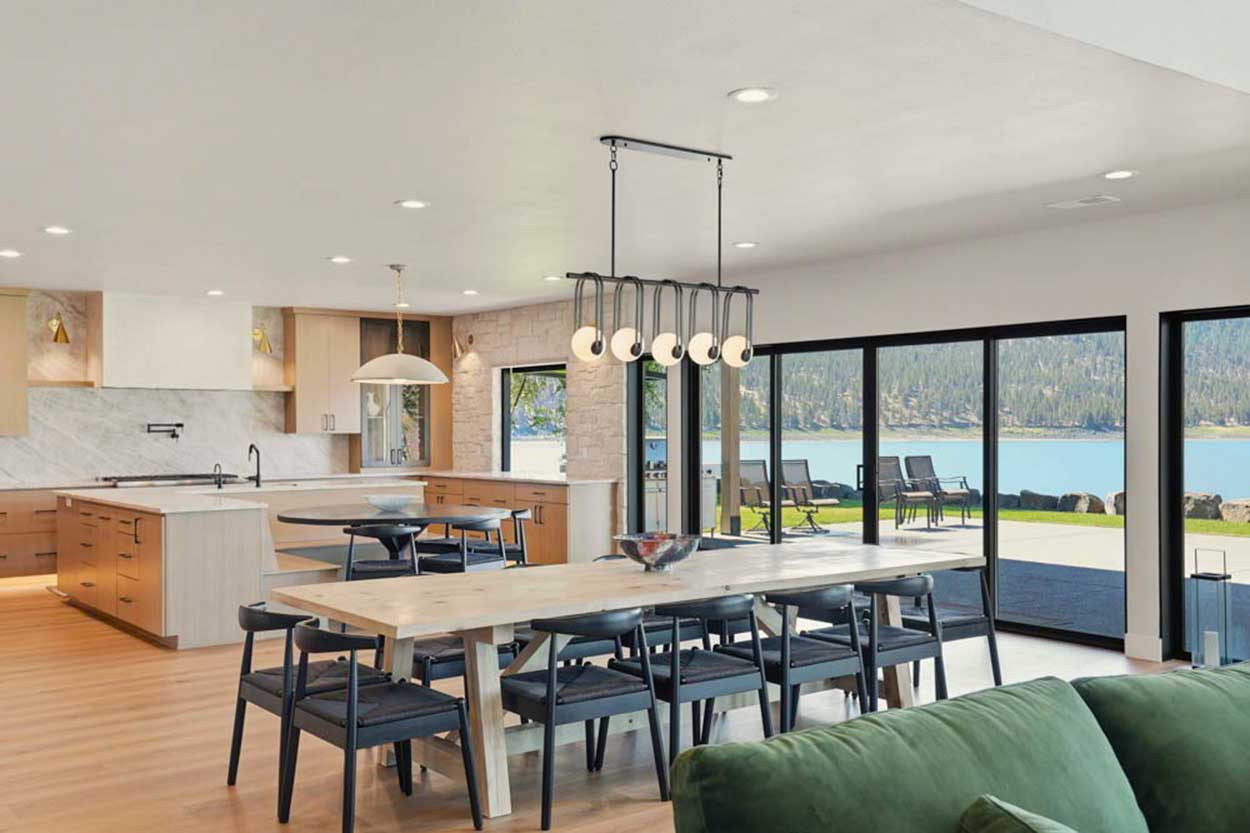 Dining area with a long whitewashed trestle table, black wishbone chairs, linear globe chandelier, and sliders to the lakeside patio.