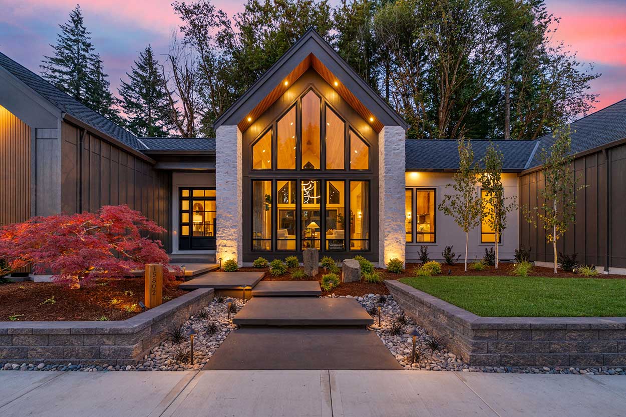Modern farmhouse exterior at dusk with a glowing two-story gabled window wall, white stone pillars, and floating concrete entry steps.