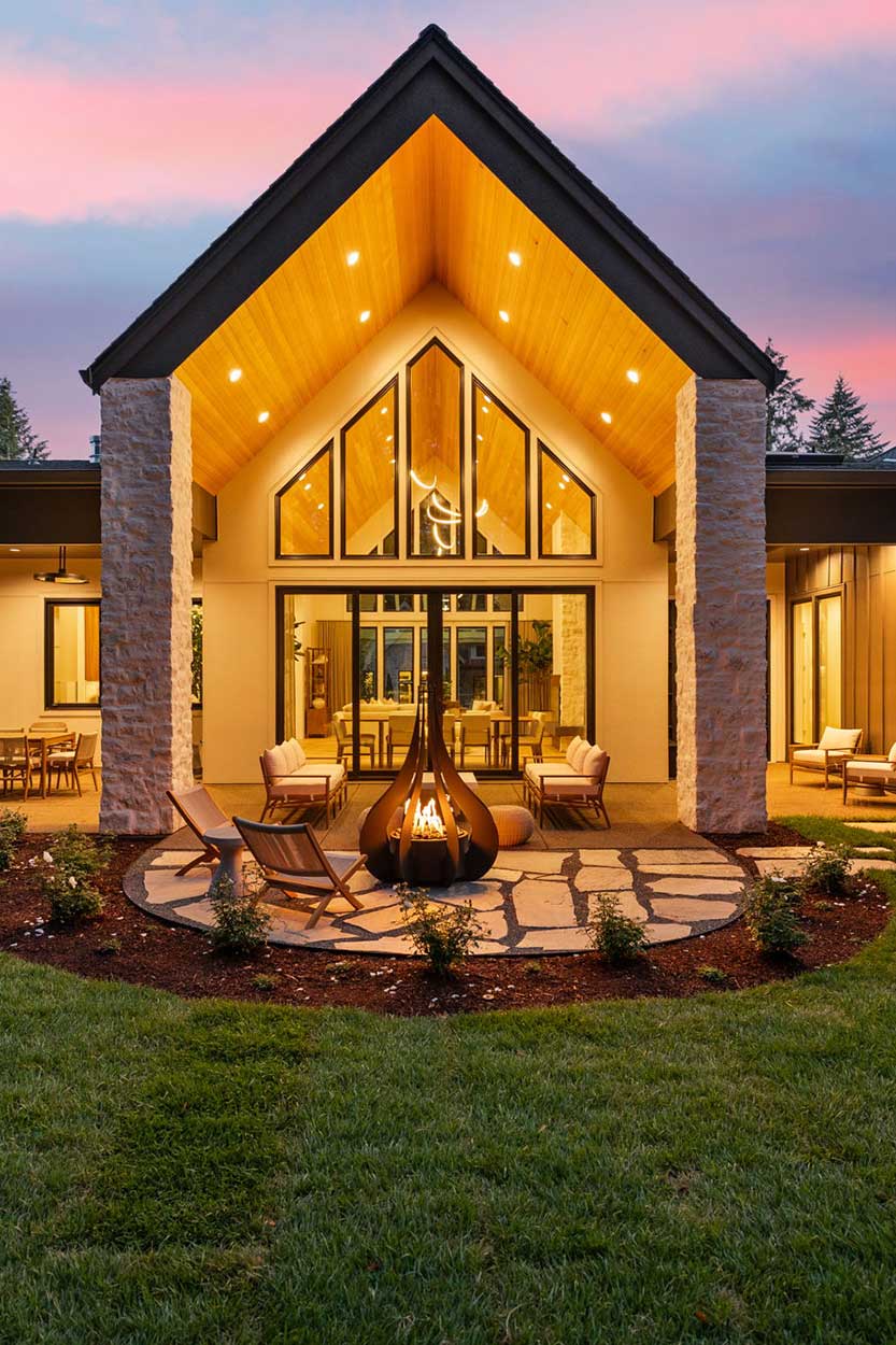 cropped view of ear courtyard at dusk with a lit fire pit, glowing gabled window wall, and outdoor kitchen and dining under covered patios.